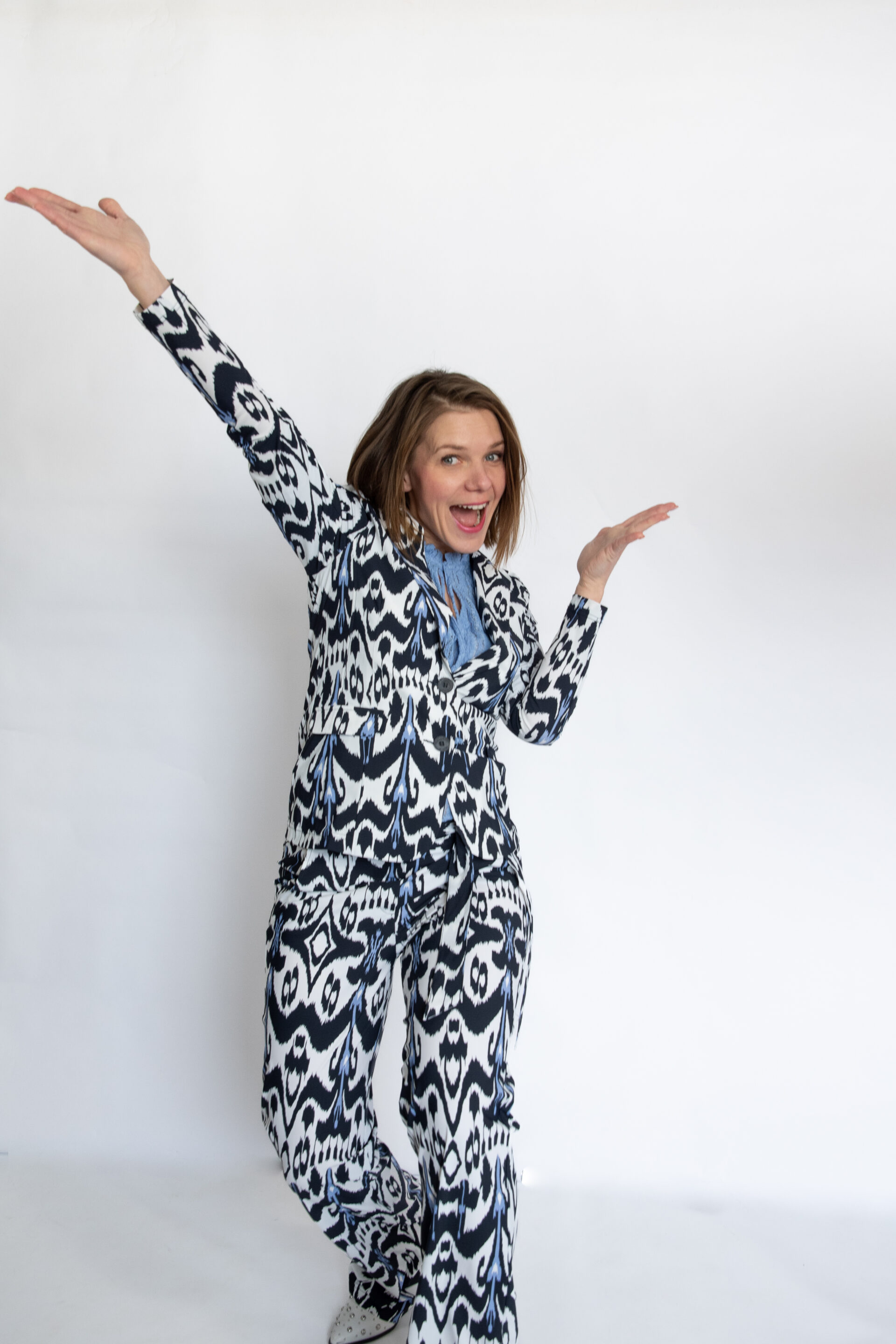 Woman in a black-and-white patterned suit raises both arms and smiles against a white background.