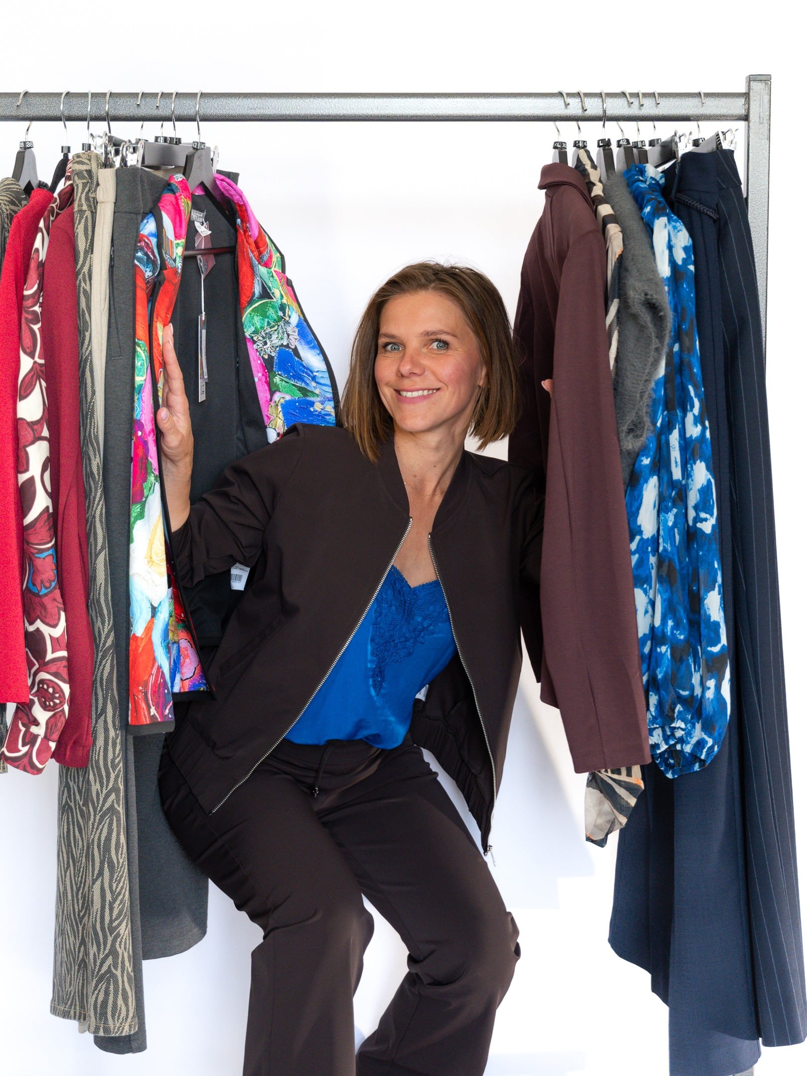 Smiling woman in a dark outfit posing between racks of colorful jackets on a clothes rail against a white background.